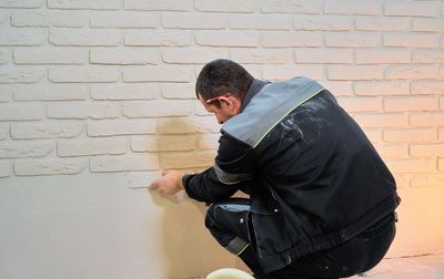 Une personne applique du plâtre sur un mur de briques blanches à l'intérieur, accroupie et travaillant avec un outil.