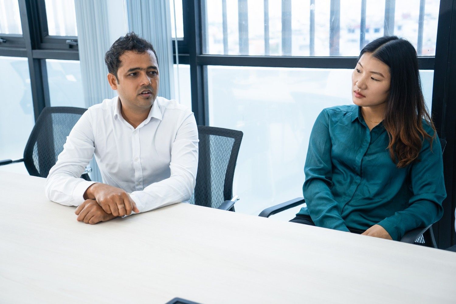 Dos personas sentadas a una mesa, mirando en la misma dirección. Oficina con ventanas.