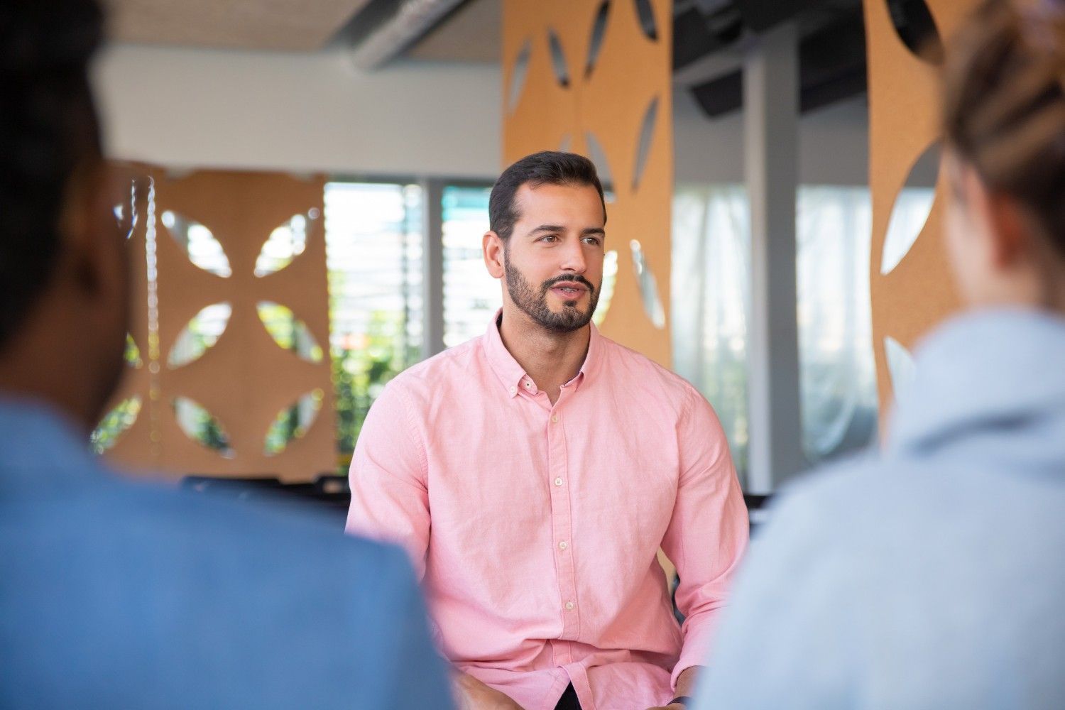 Un hombre con camisa rosa se sienta en círculo, mirando a otras personas; entorno de oficina con pared decorativa.