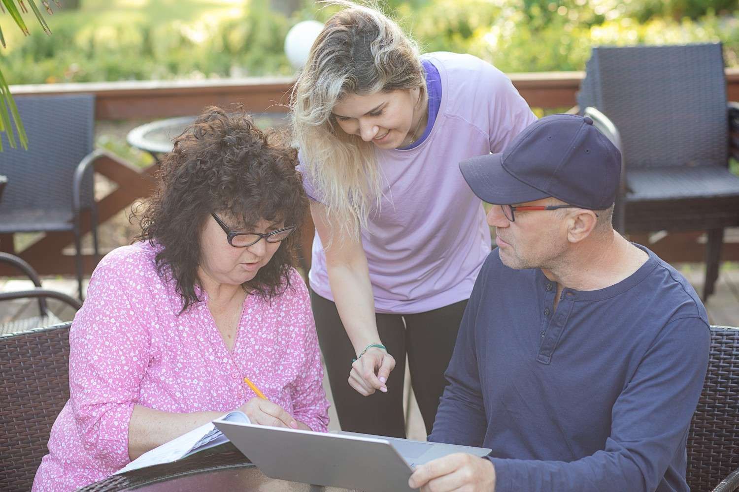 Tres personas revisan documentos en una mesa al aire libre; una mujer escribe y otras dos observan.