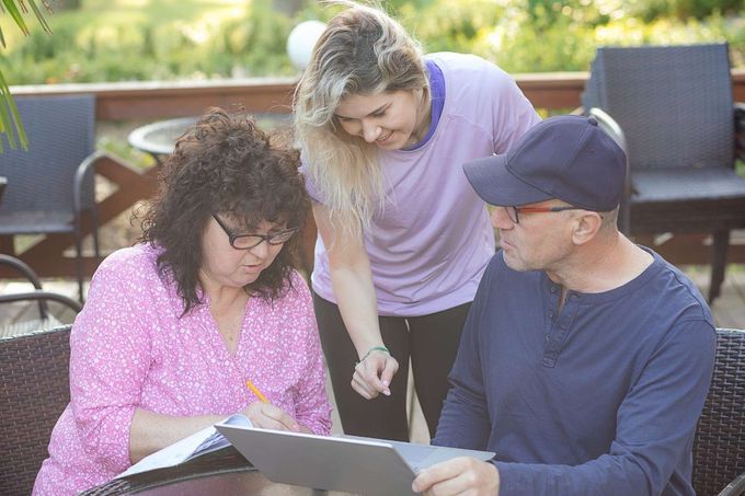 Tres personas revisan documentos en una mesa al aire libre; una mujer escribe y otras dos observan.