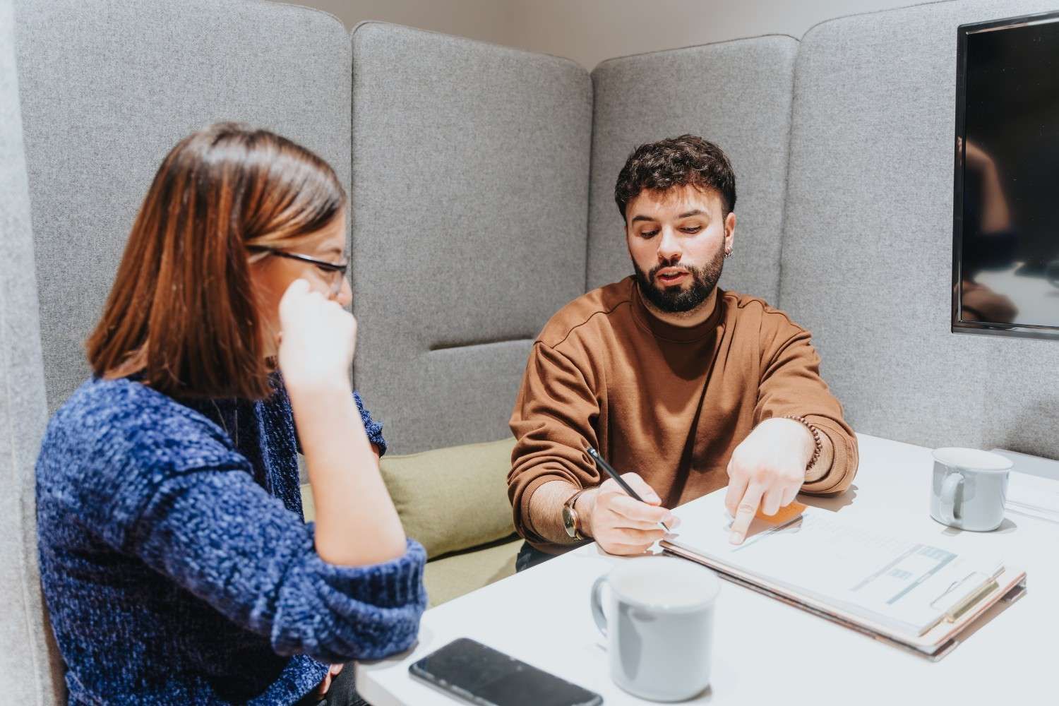 Mujer y hombre en una reunión, hombre escribiendo en un bloc de notas, tazas y un teléfono sobre la mesa de una cabina.