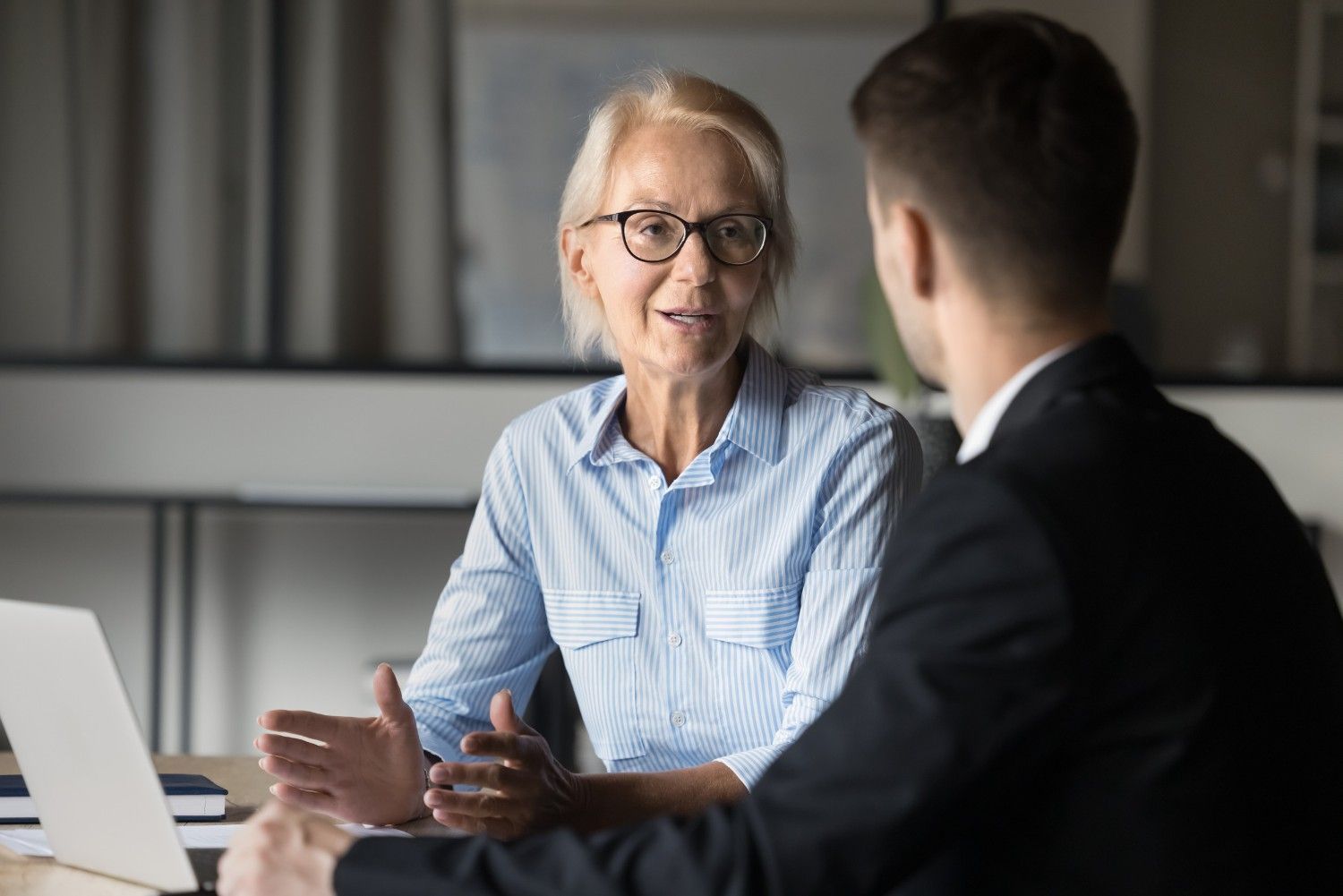 Una mujer con gafas hace gestos mientras habla con un hombre de traje, ambos sentados en un escritorio con una computadora portátil.