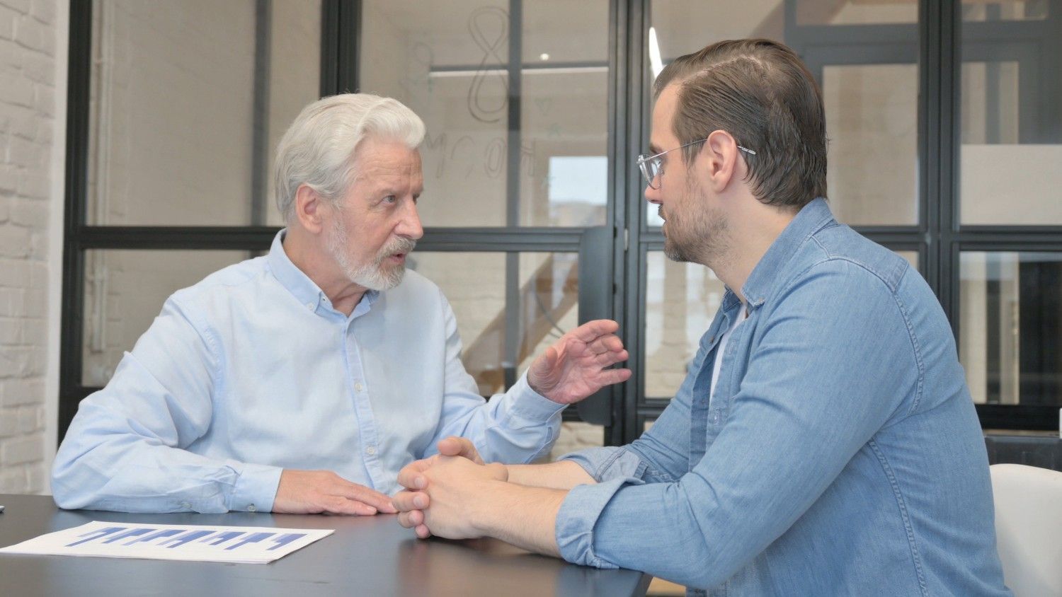 Dos hombres sentados a una mesa, gesticulando y hablando con expresiones serias; gráficos sobre la mesa.