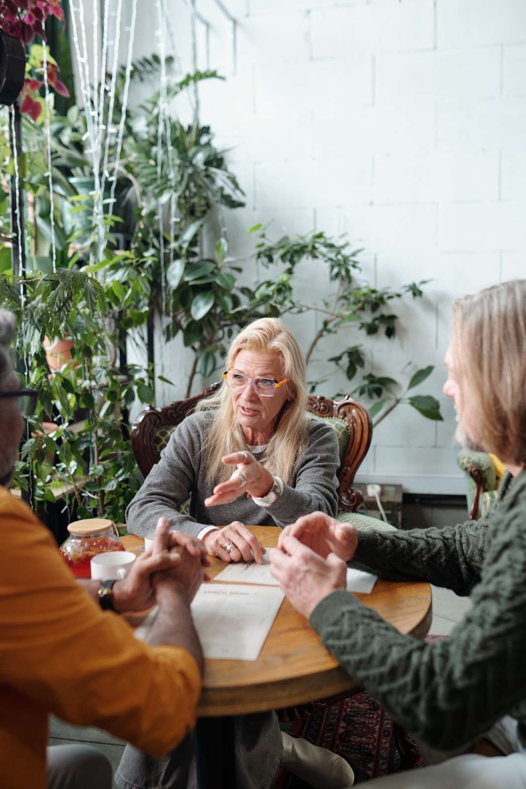 Tres personas sentadas alrededor de una mesa, una de ellas gesticulando con las manos, con plantas en el fondo.