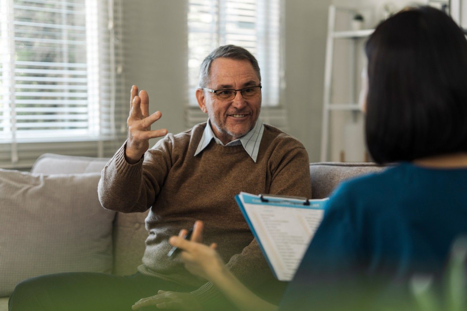 Un hombre con gafas gesticula mientras habla con alguien que sostiene un portapapeles. Están sentados en una sala de estar.