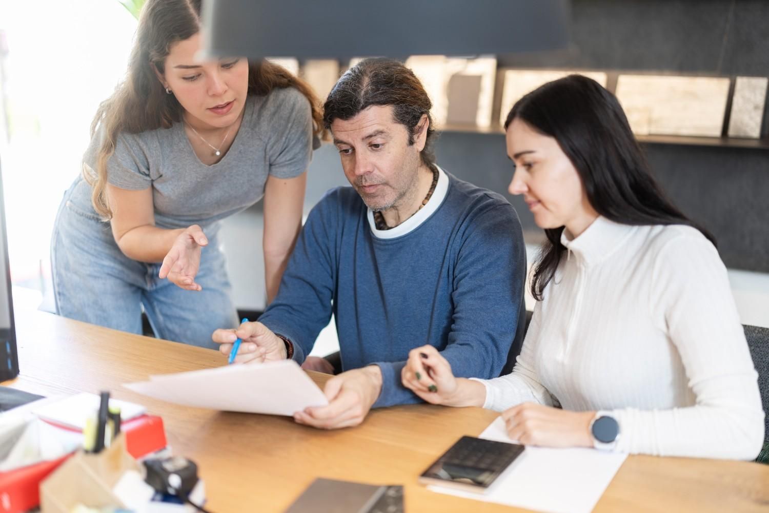 Tres personas revisando documentos en una mesa. Dos mujeres y un hombre. Interior.