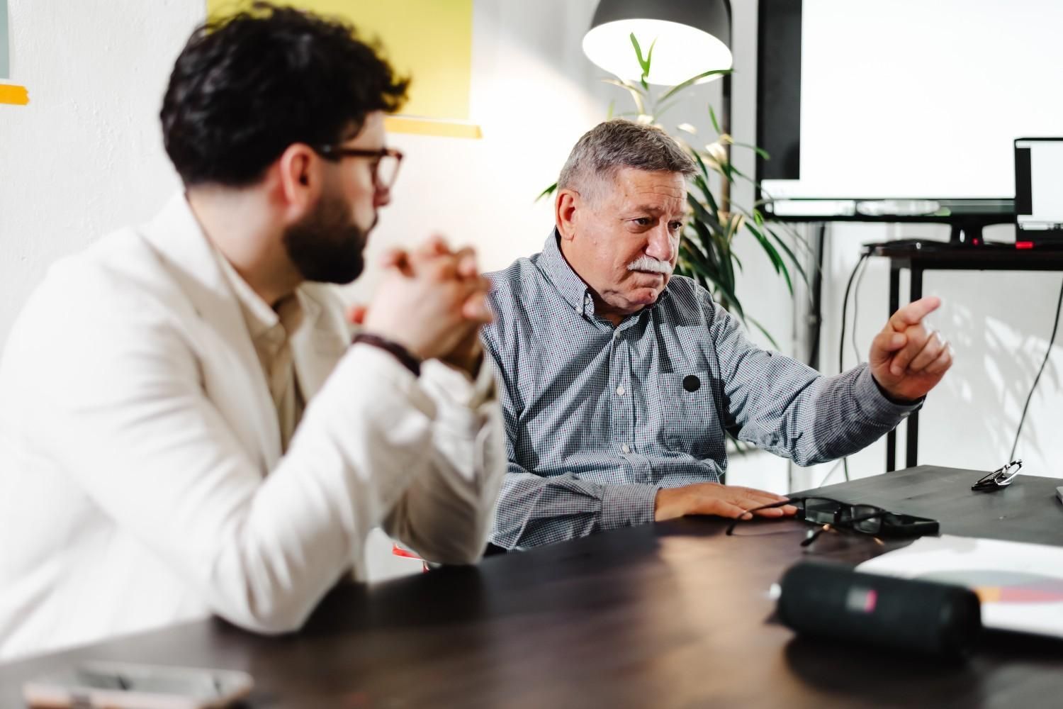 Dos hombres en una mesa, uno señalando, discutiendo documentos, en un entorno de oficina.
