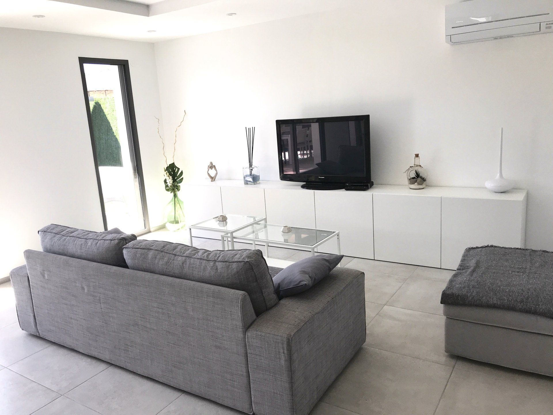 Salon moderne avec canapés gris, table basse en verre, télévision et meubles blancs.