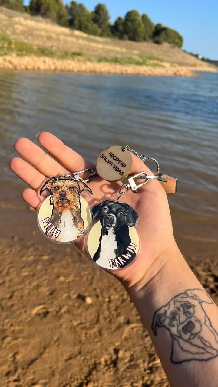 Mano sosteniendo tres llaveros de perro y una etiqueta de madera, con un tatuaje de perro en el brazo, fondo de lago.