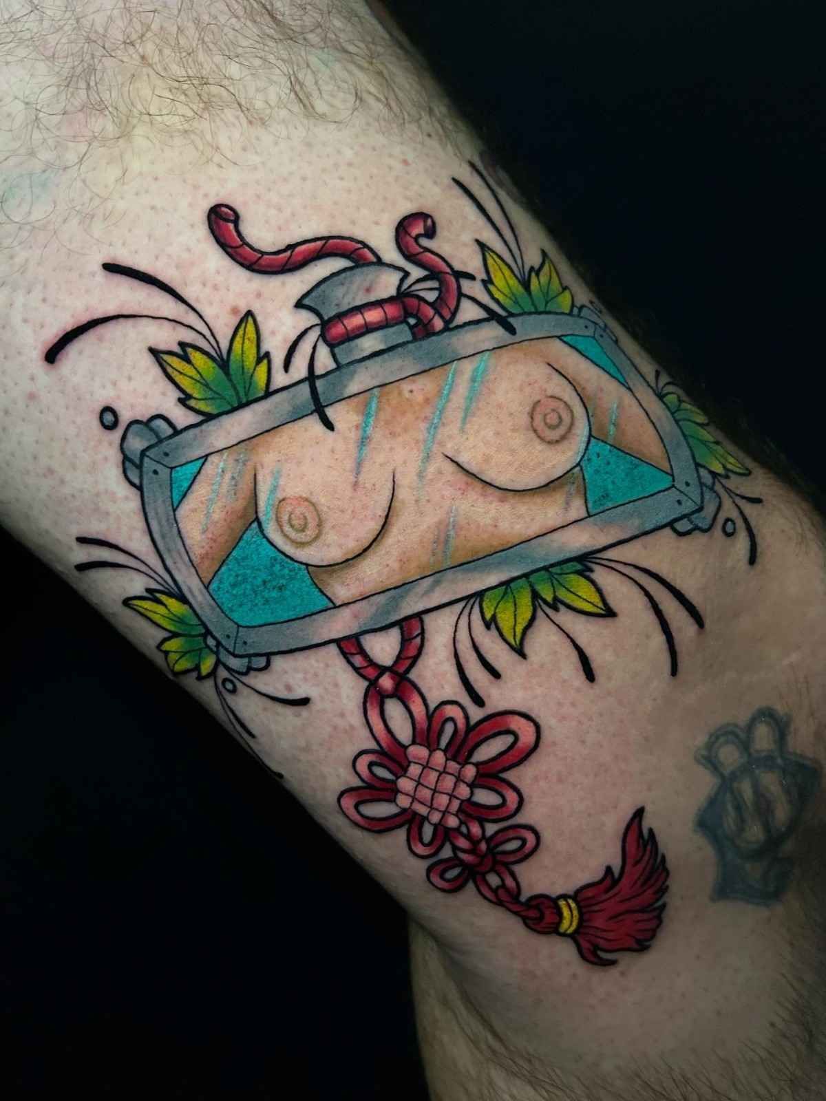 Tatuaje de un espejo retrovisor que refleja los pechos de una mujer, con un nudo rojo y acentos florales.