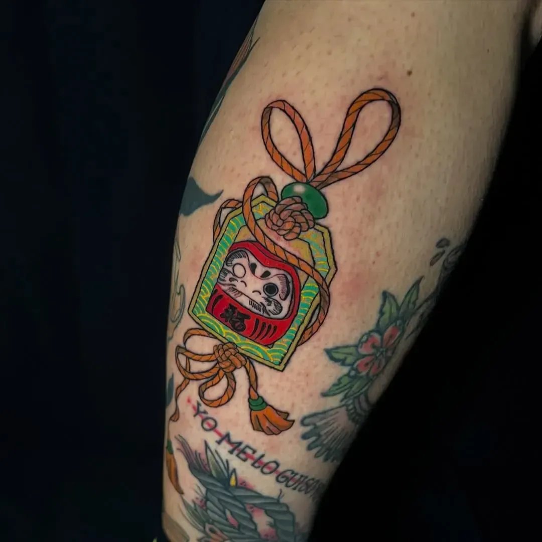 Tatuaje de un muñeco Daruma japonés en una bolsa verde con dijes y cuerda naranja. Tatuaje en el brazo.
