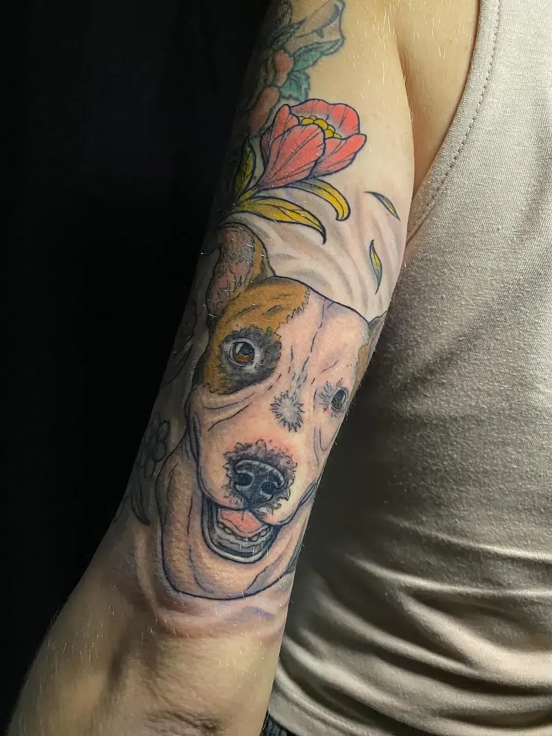 Tatuaje de la cara sonriente de un perro con una flor. El perro tiene pelaje marrón y blanco sobre un fondo negro.