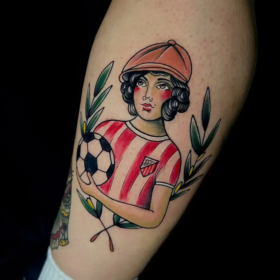 Tatuaje de una persona con camiseta y gorra de rayas rojas, sosteniendo un balón de fútbol. Hojas verdes rodean la figura.