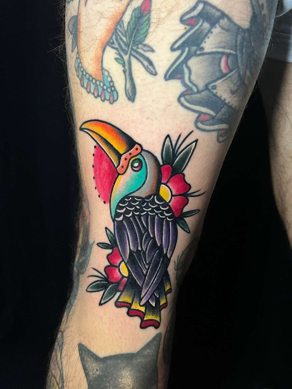 Tatuaje de un tucán con pico y plumas de colores, rodeado de flores rojas.