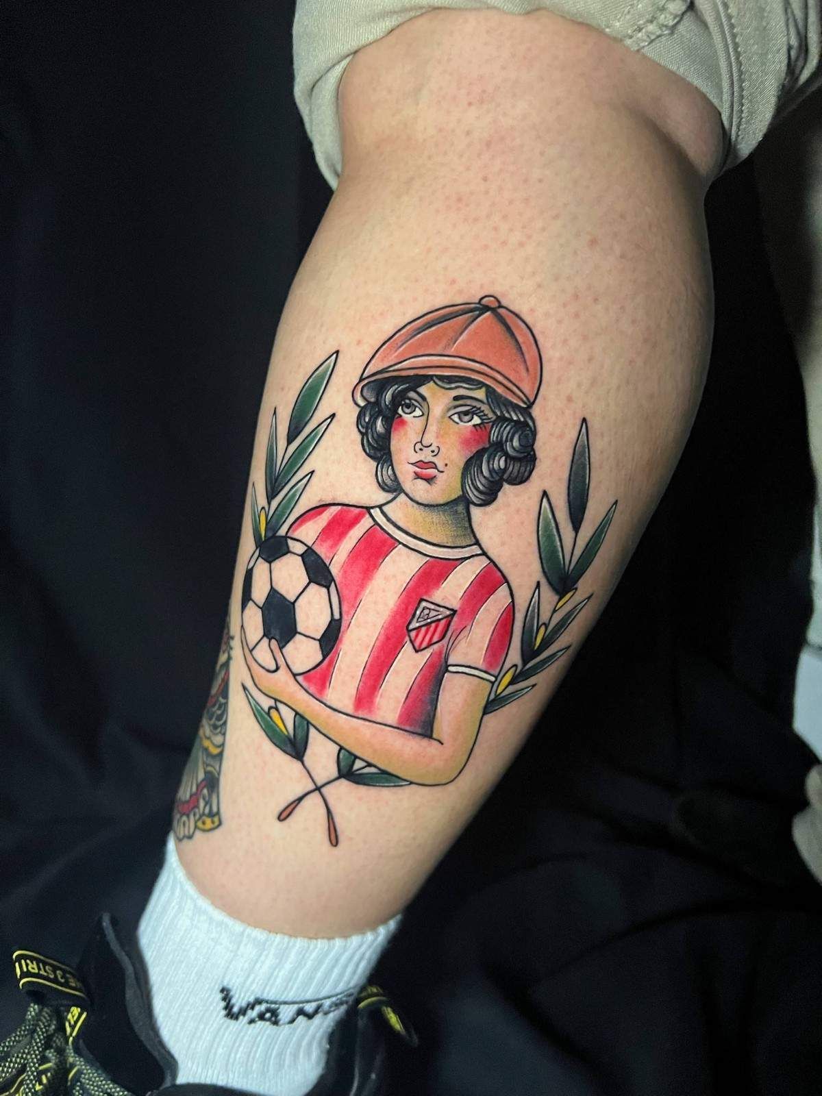 Tatuaje de una persona con camiseta y gorra de rayas rojas y blancas, sosteniendo un balón de fútbol, rodeado de hojas.