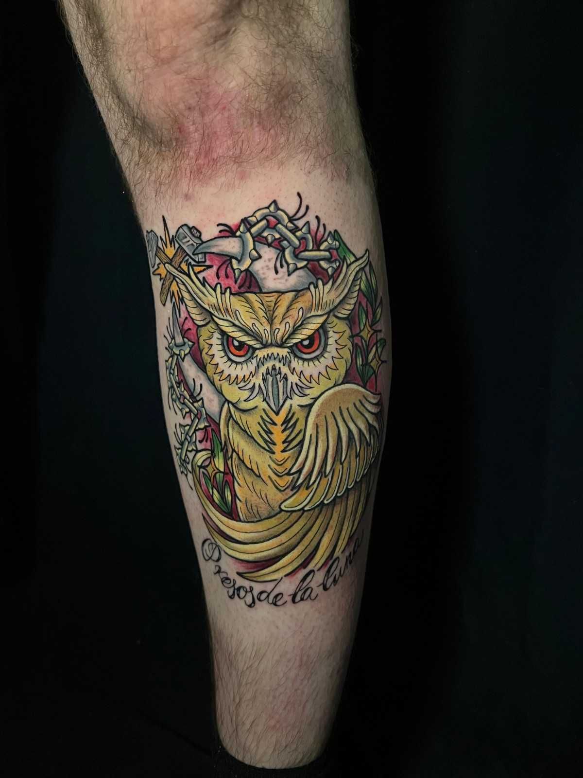 Tatuaje de un búho con ojos rojos, rodeado de flores y una pancarta que dice