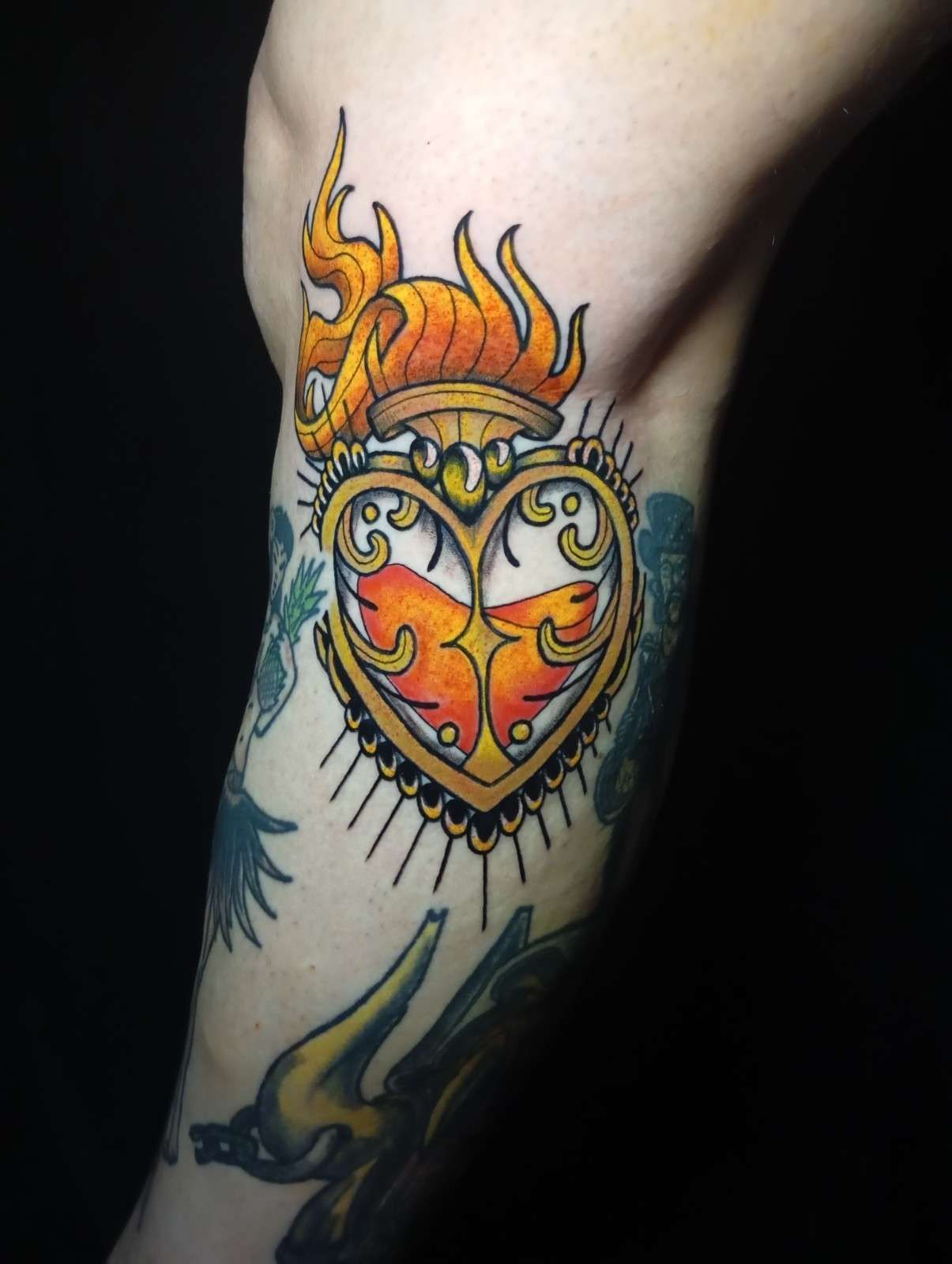 Tatuaje de un frasco dorado en forma de corazón con llamas en un brazo, rodeado de otros tatuajes.