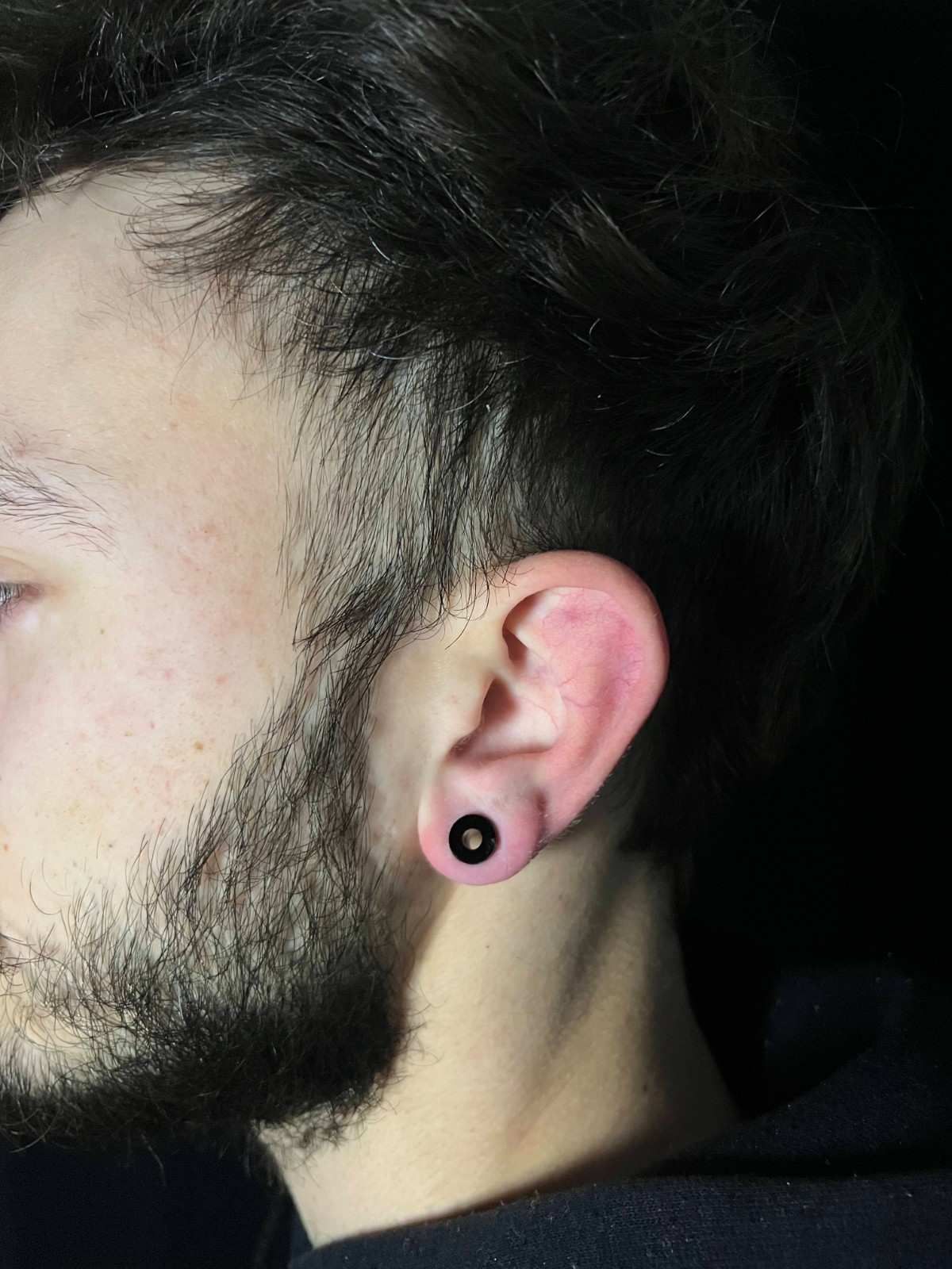 Oreja de hombre con tapón negro; barba oscura, cabello oscuro, perfil lateral.