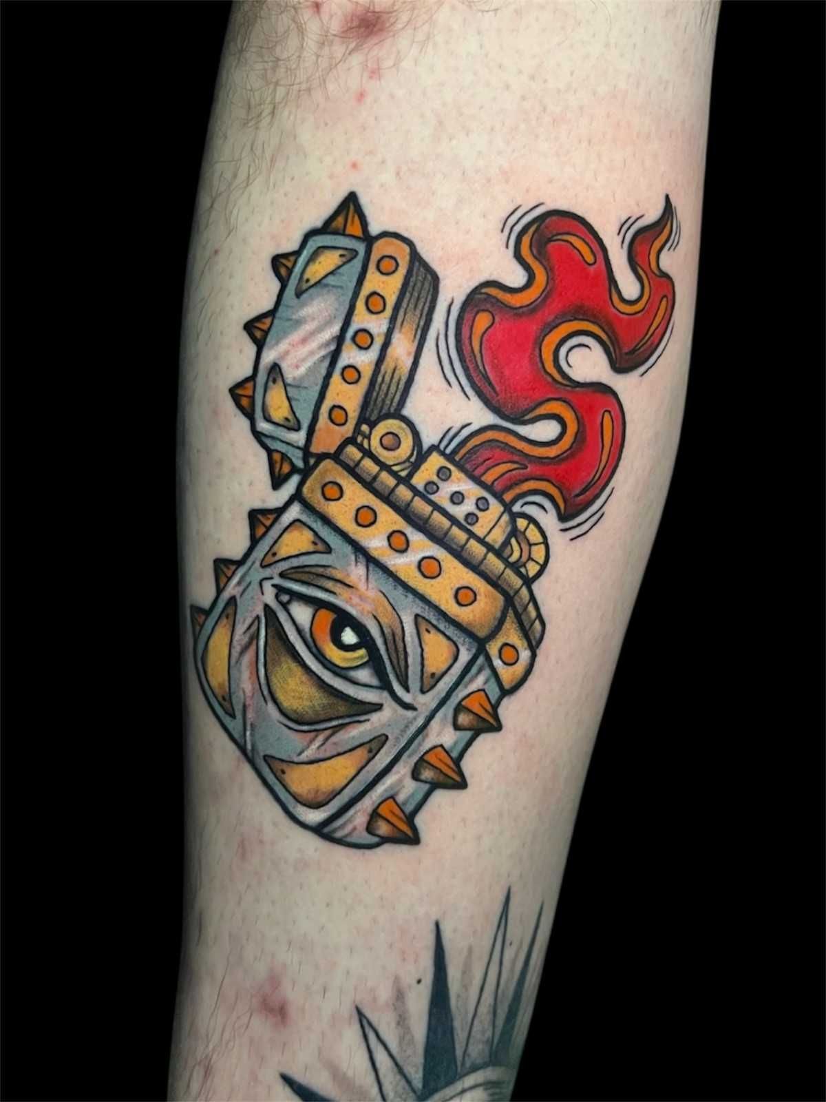 Tatuaje de un encendedor estilizado con un ojo. Tiene llamas y púas, en tonos grises, amarillos y rojos.