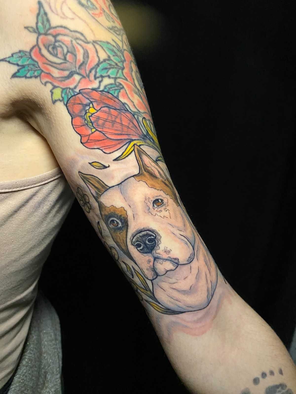 Tatuaje en el brazo que presenta un retrato de perro con flores en rojo, rosa y verde sobre un fondo oscuro.