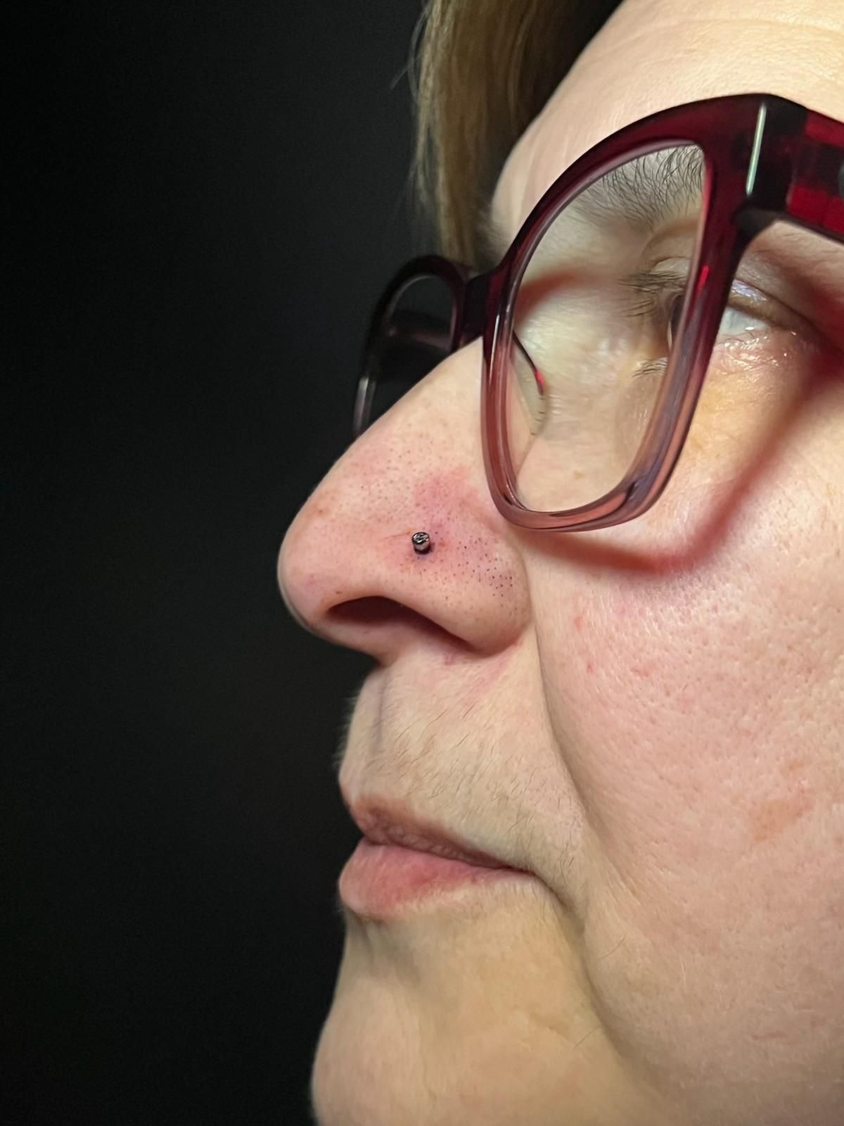Mujer con piercing en la nariz y gafas, vista de perfil. Fondo oscuro.