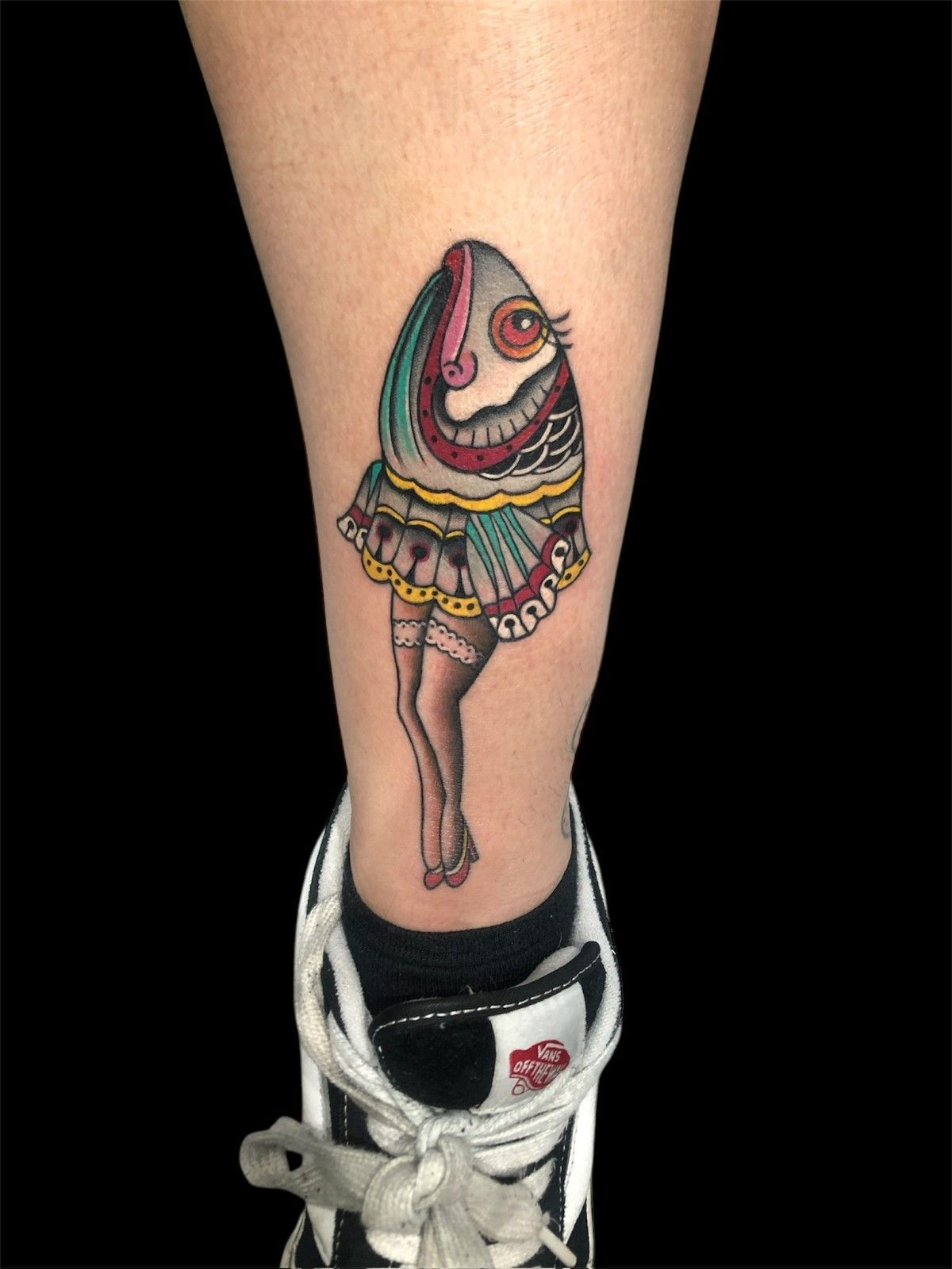 Tatuaje de una figura estilizada y caricaturesca con forma de huevo, con piernas y falda, en una pierna