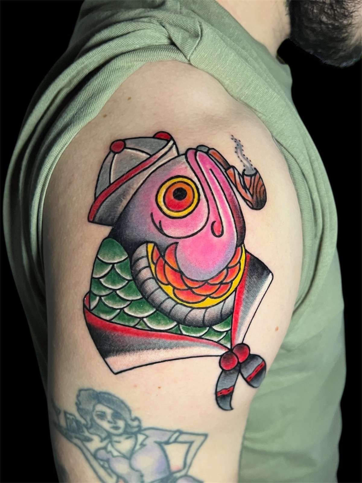 Tatuaje de un pez con sombrero y fumando una pipa, en la parte superior del brazo de una persona