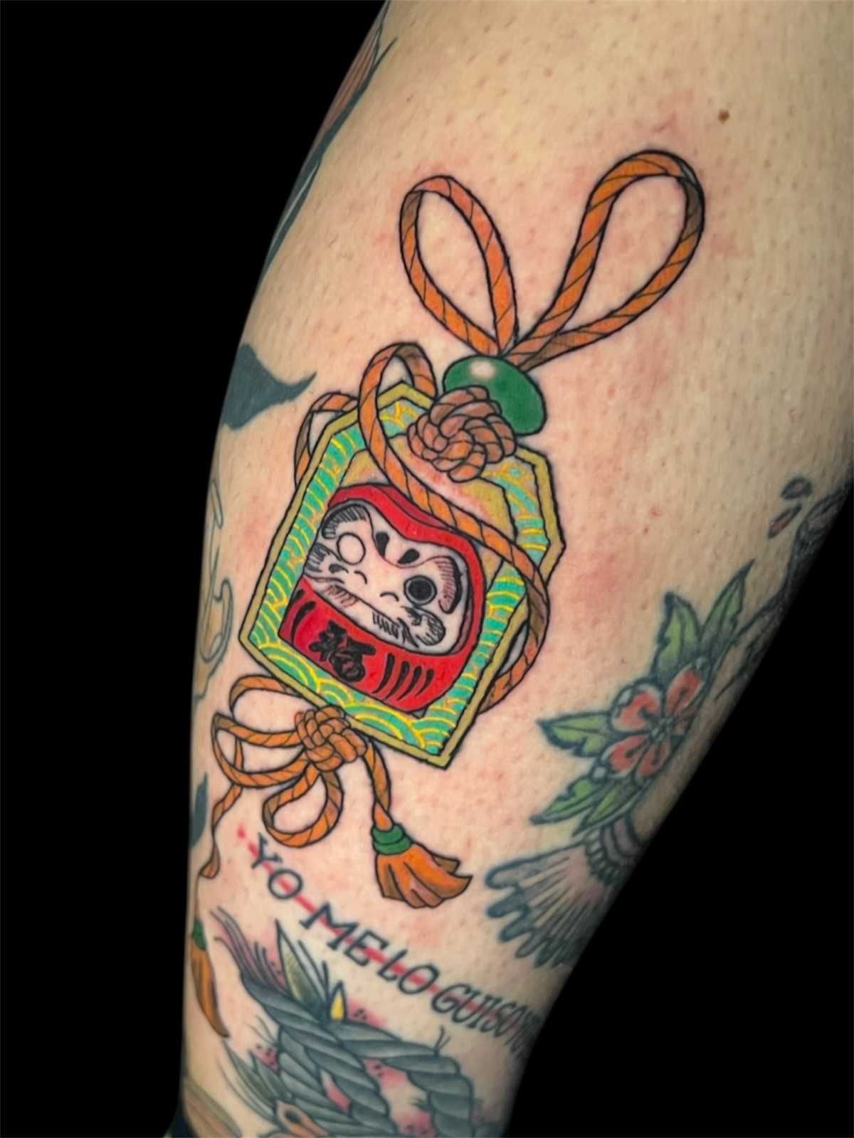 Tatuaje de un dije de muñeca Daruma en un antebrazo; rojo, blanco y verde con un cordón naranja anudado.