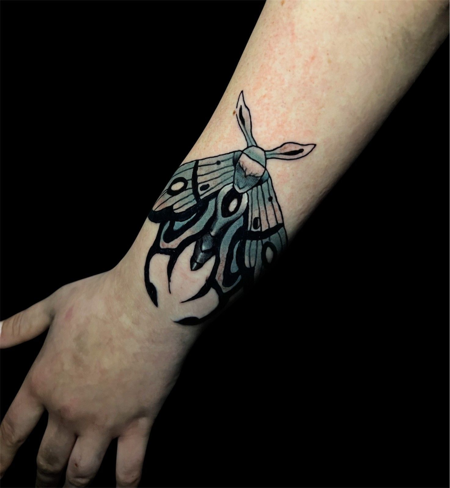 Tatuaje de una polilla azul estilizada en una muñeca con diseños negros similares a llamas debajo.