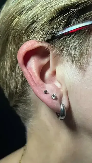 Oreja con tres piercings: dos pequeños y un aro de plata. Cabello rubio visible.