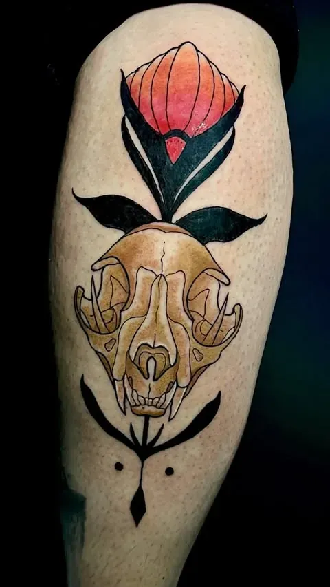 Tatuaje de una calavera de gato con una flor estilizada y hojas en negro, rojo y beige sobre la piel.