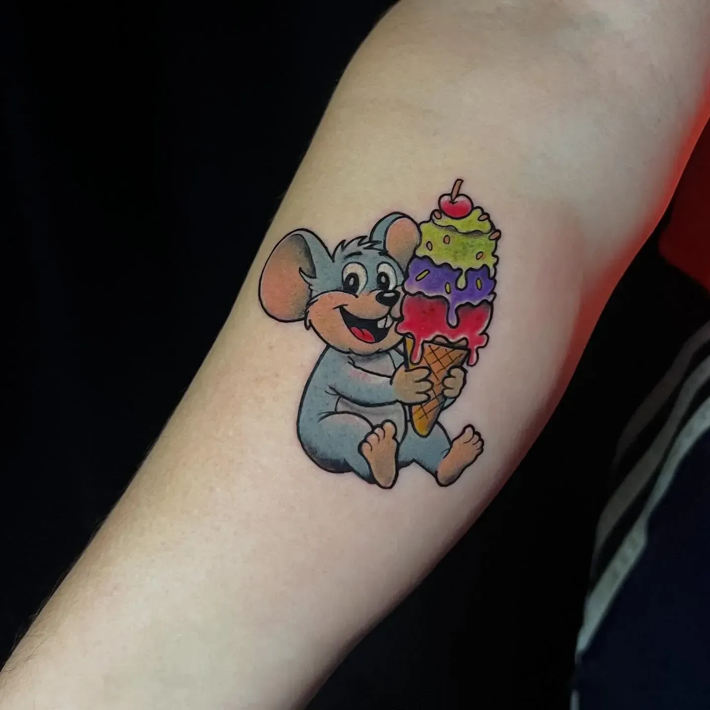 Tatuaje de un ratón de dibujos animados que sostiene un colorido cono de helado de tres bolas con una cereza encima.