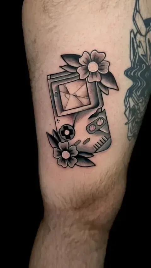 Tatuaje en color negro y gris de un Game Boy roto con flores en una pierna.