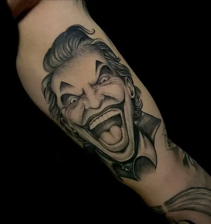 Tatuaje de la cara sonriente del Joker en el brazo de una persona, sombreado negro y gris.