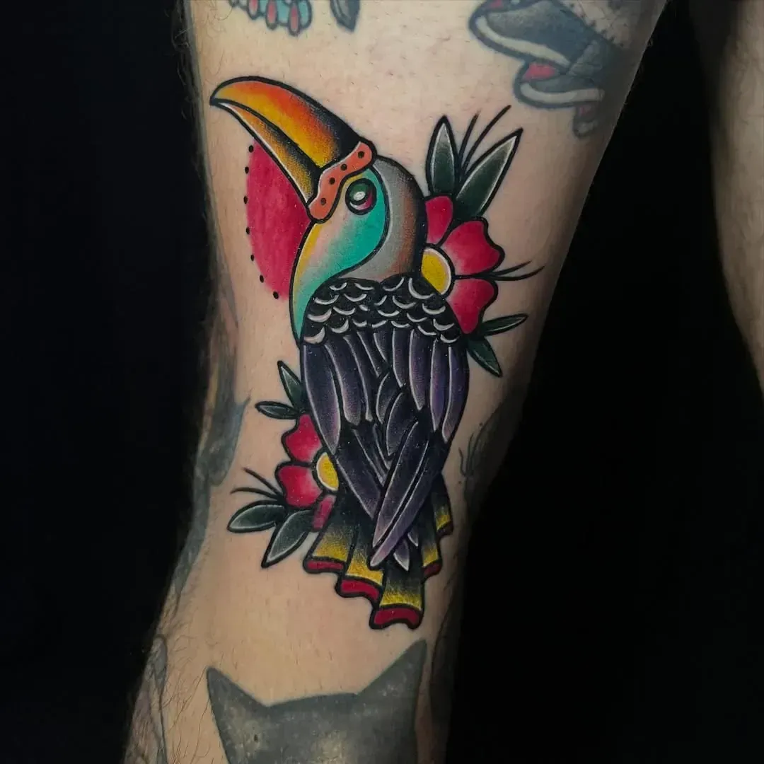 Tatuaje tradicional de un tucán colorido en una rodilla, rodeado de flores rojas y hojas verdes.