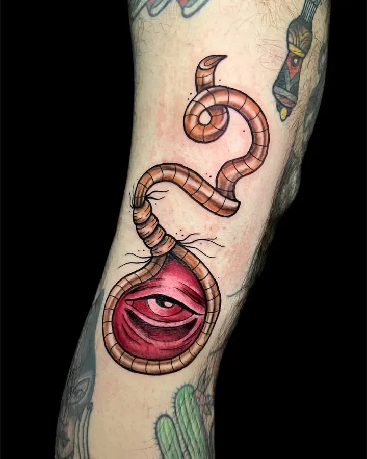 Tatuaje en la pierna: ojo rojo dentro de un diseño marrón similar a un gusano; otros tatuajes visibles.