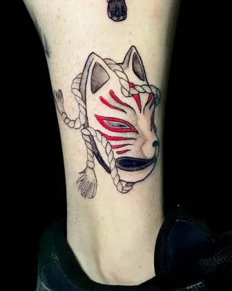 Tatuaje de una máscara kitsune blanca con marcas rojas y detalle de cuerda en una pierna. Fondo negro.