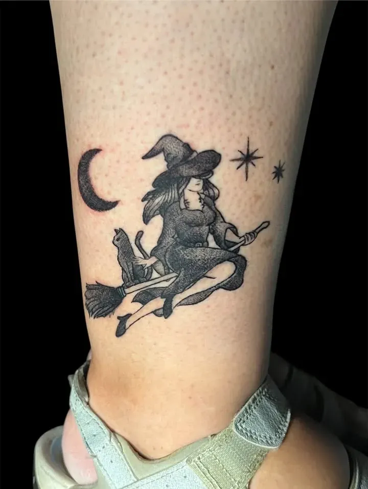 Tatuaje de una bruja volando en una escoba con un gato negro, una luna creciente y estrellas en un tobillo.