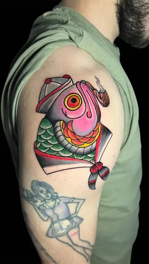 Tatuaje de un pez con sombrero y fumando en pipa. Está en la parte superior del brazo.
