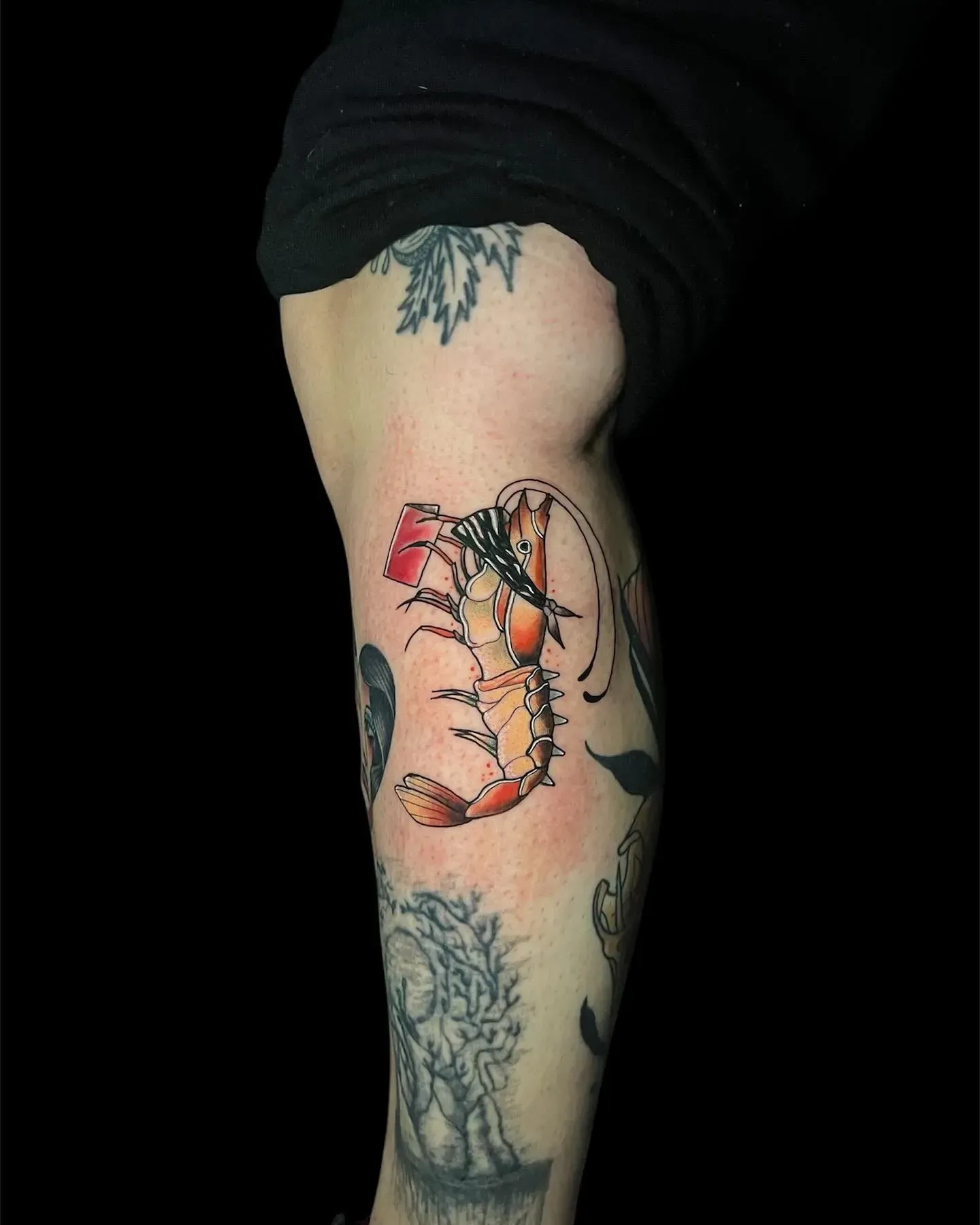 Tatuaje en la pierna de un camarón colorido con sombrero. También hay otros tatuajes.