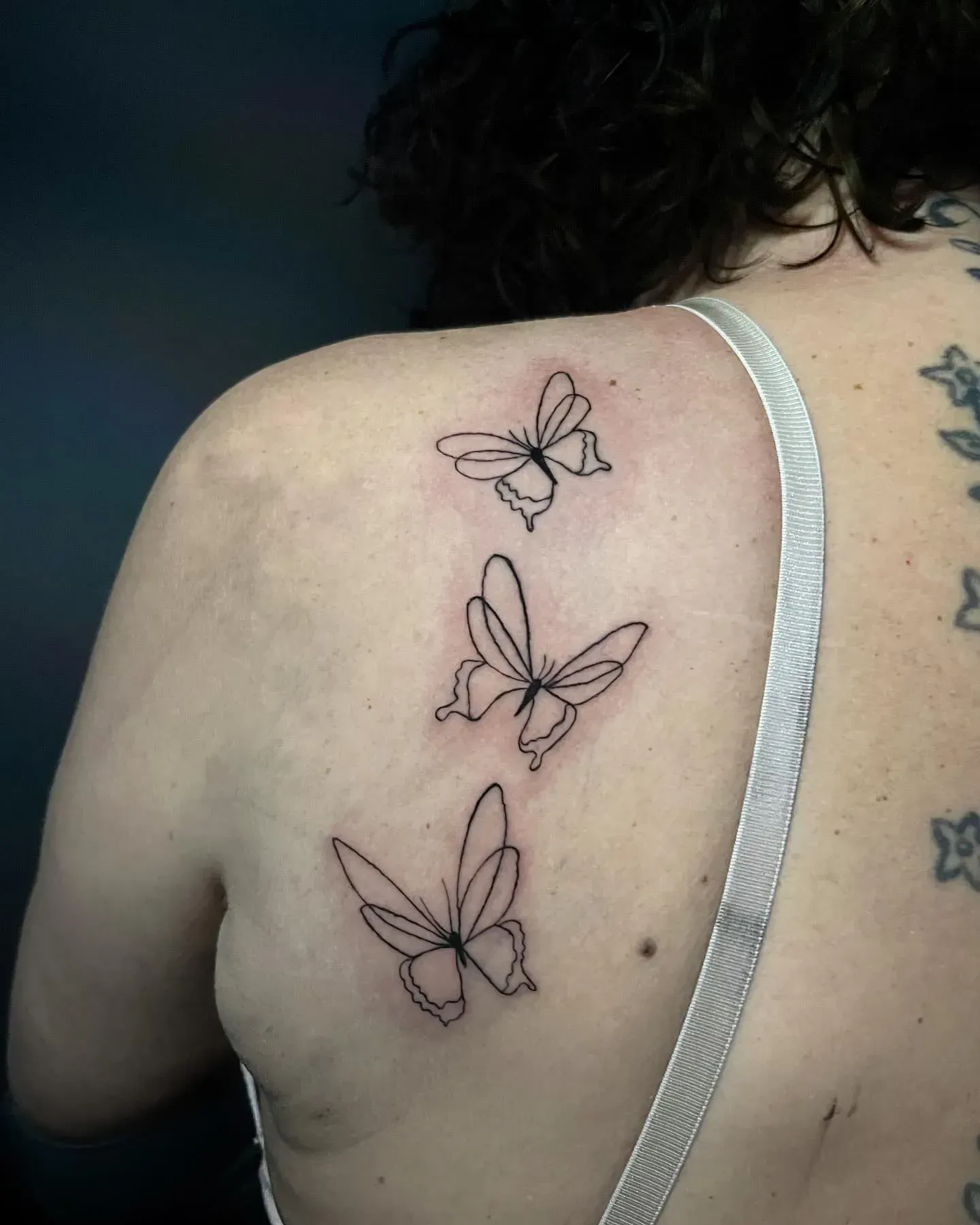 Tres pequeños tatuajes de mariposas de contorno negro en la espalda de una persona, cerca del hombro.
