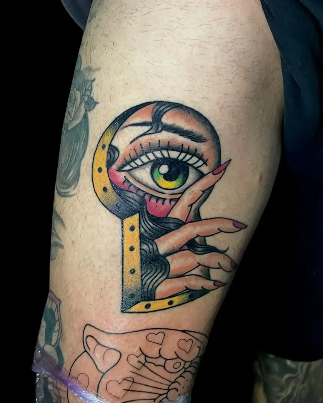 Tatuaje de un ojo asomándose por el ojo de una cerradura, con una mano sosteniendo un dedo hacia arriba