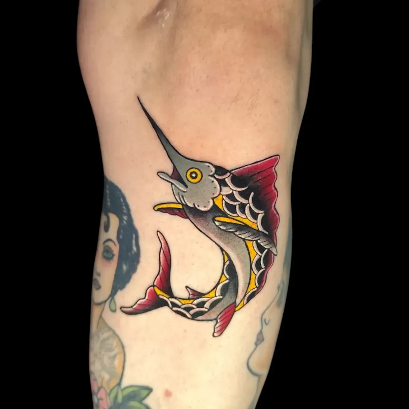 Tatuaje de un pez espada en estilo tradicional sobre piel, con colores rojo, amarillo y negro.