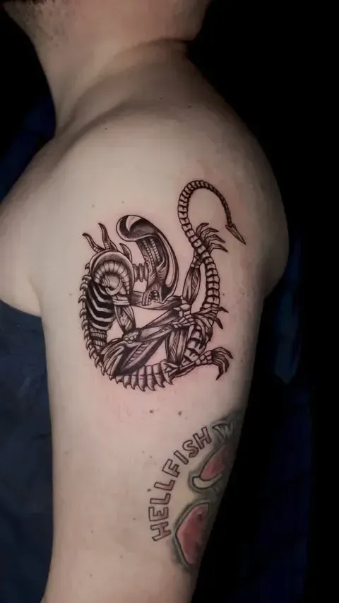 Tatuaje de un extraterrestre xenomorfo en la parte superior del brazo de una persona, con detalles esqueléticos y una cola.