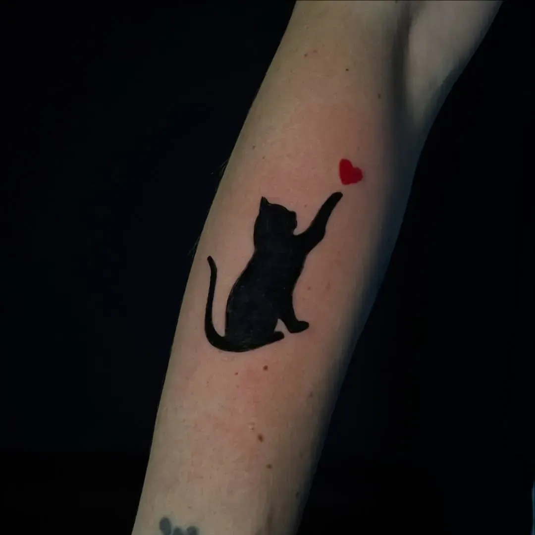 Silueta de gato negro que alcanza un tatuaje de corazón rojo en un brazo.