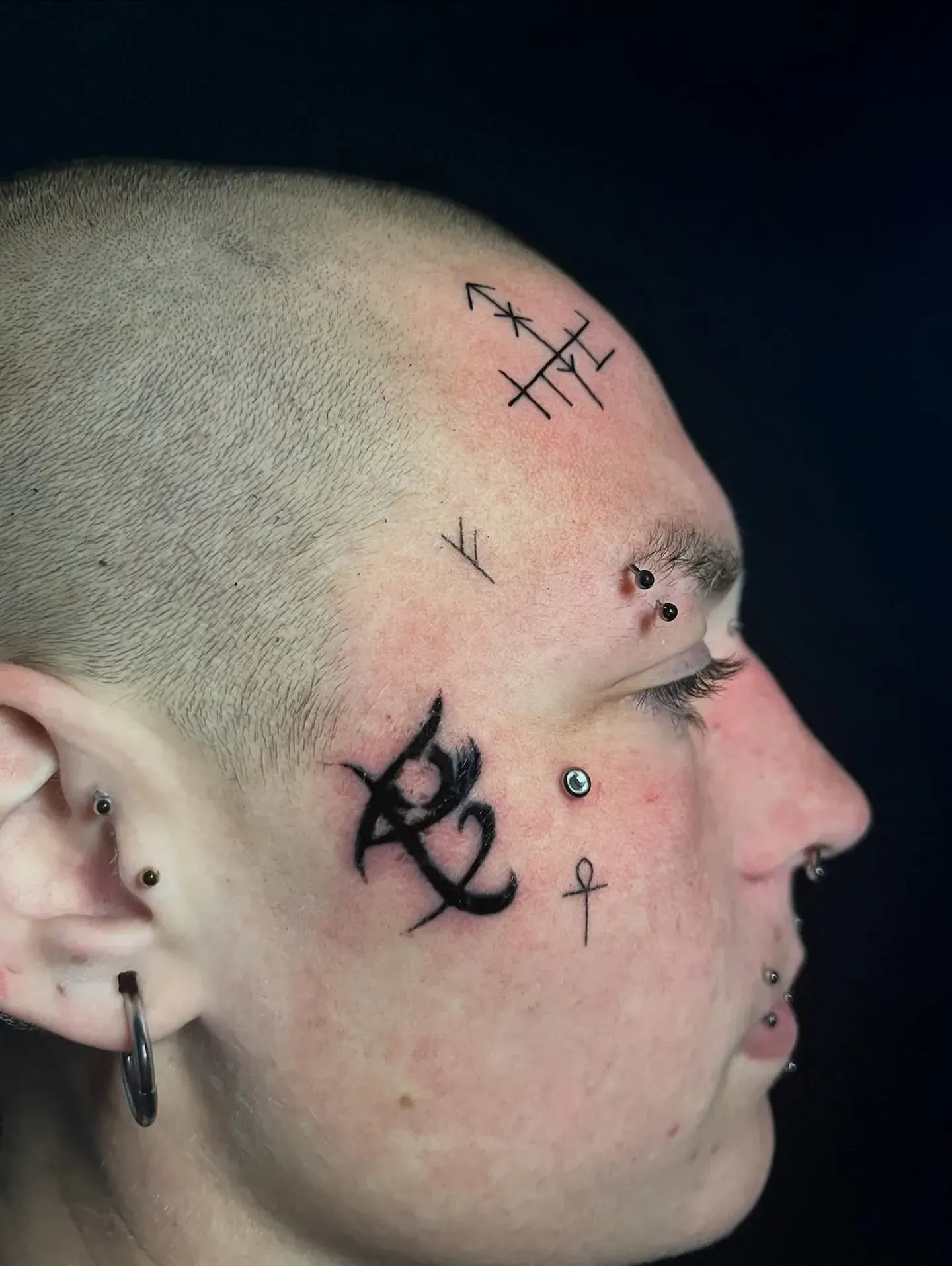 Persona con tatuajes en la cara, piercings y cabeza rapada, de perfil.