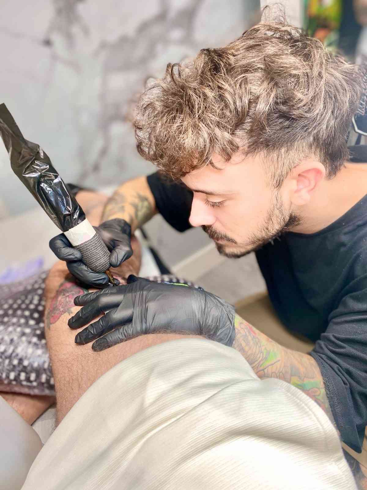 Hombre con cabello rizado y barba, con los brazos cruzados, vistiendo una chaqueta negra y verde azulado