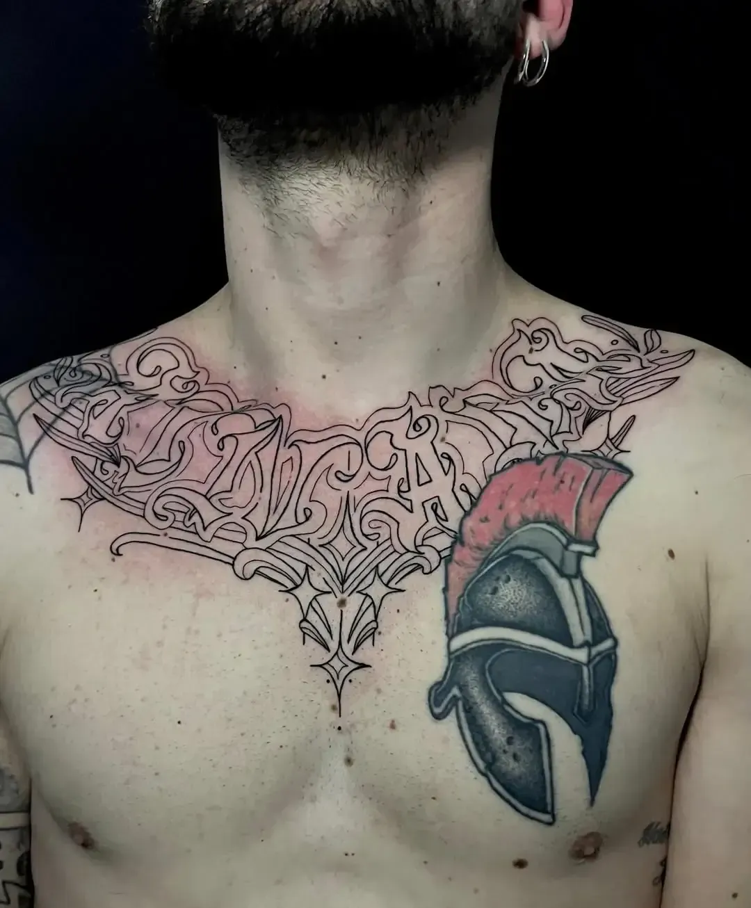 Tatuaje en el pecho de un hombre: diseño intrincado alrededor del cuello, casco espartano en el lado derecho.