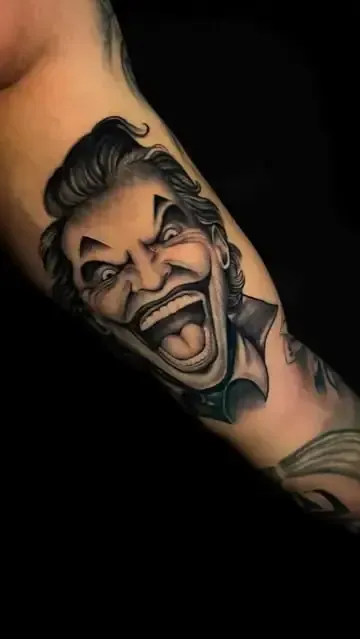 Tatuaje del Joker sonriendo ampliamente, con dientes afilados y ojos salvajes, en el brazo de una persona